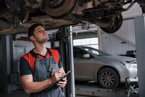 Counting how many details needed. Man at the workshop in uniform using notepad for his job for fixing broken car.