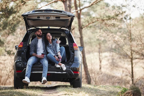 休憩中だ 車の後部に座っている 自然を楽しむ 夫婦は彼らのブランドの新しい黒い車で森に到着しました — ストック写真