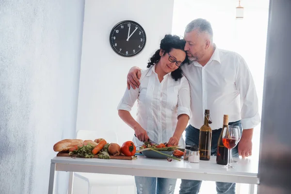 敏感な抱擁 台所で野菜を使った料理を作る白いシャツを着た男と妻 — ストック写真