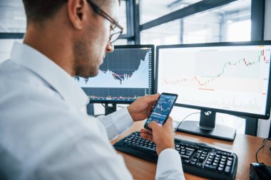 Akıllı telefon kullanıyor. Resmi giyinmiş genç iş adamı birden fazla ekranla ofiste. Değişim ve para kavramı.