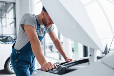 Hiç fena görünmüyor. Mavi üniformalı ve siyah şapkalı adam hasarlı otomobili tamir ediyor..