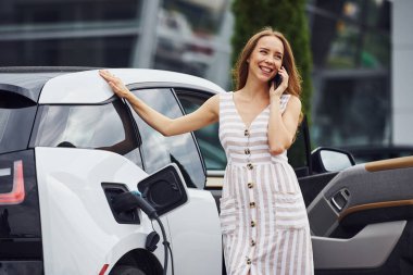 Modern kablosuz teknolojiler. Telefonla. Elektrikli arabalardaki kadın gündüz şarj istasyonunda. Yepyeni bir araç..
