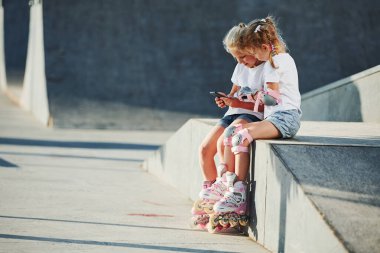 Akıllı telefon kullanıyor. Ekstrem sporlar için rampada. Dışarıda patenli iki küçük kız eğleniyor..