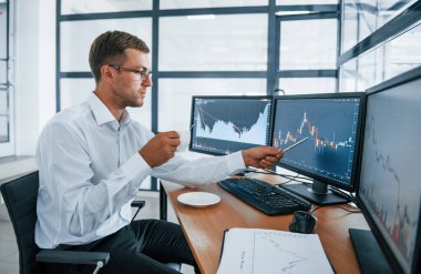 Bir bardak içki tutuyor. Bilgi analiz ediliyor. Resmi giyinmiş genç iş adamı birden fazla ekranla ofiste. Değişim ve para kavramı.