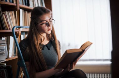 Oturup yeni bilgiler öğreniyor. Kütüphanede kitaplarla dolu bir kız öğrenci var. Eğitim kavramı.