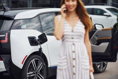 Resim odaklama tekniği. Telefonla. Elektrikli arabalardaki kadın gündüz şarj istasyonunda. Yepyeni bir araç..