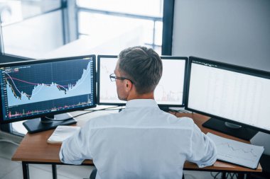 Meşgul bir çalışan. Arkadan bak. Resmi giyinmiş genç iş adamı birden fazla ekranla ofiste. Değişim ve para kavramı.