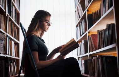 Oturup yeni bilgiler öğreniyor. Kütüphanede kitaplarla dolu bir kız öğrenci var. Eğitim kavramı.