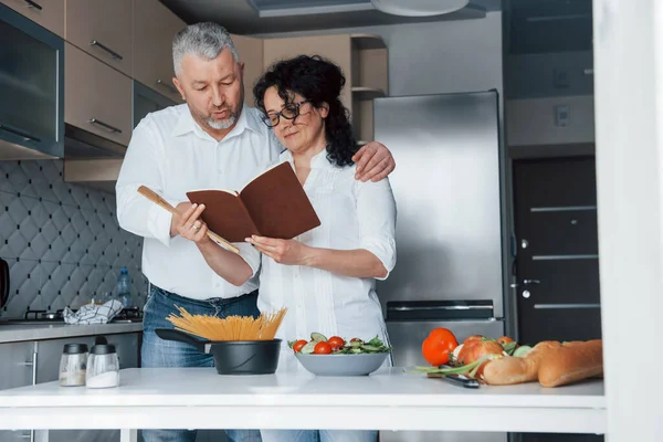 本を読んでいる 台所で野菜を使った料理を作る白いシャツを着た男と妻 — ストック写真