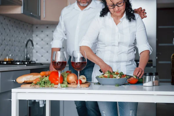 ナイフを使ってキュウリをスライスする 台所で野菜を使った料理を作る白いシャツを着た男と妻 — ストック写真