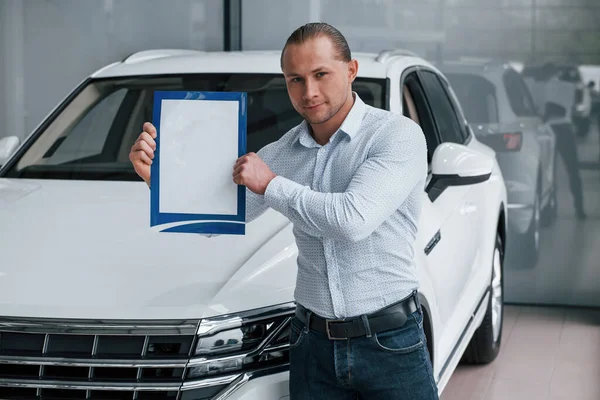 Put your text here. Manager stands in front of modern white car with paper and documents in hands.