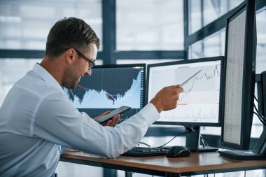 Telefon ve kalemle. Grafikler analiz ediliyor. Resmi giyinmiş genç iş adamı birden fazla ekranla ofiste. Değişim ve para kavramı.