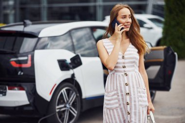 Şarj beklerken telefonu kullanmak. Gündüz vakti istasyonda bir kadın. Yepyeni bir araç..