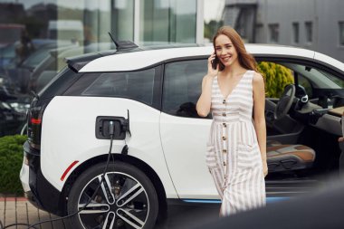 Telefon kullanarak sohbet ediyorlar. Elektrikli arabalardaki kadın gündüz şarj istasyonunda. Yepyeni bir araç..