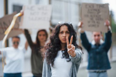 İki parmağınla jest yapıyorsun. Bir grup feminist kadın dışarıda hakları için protesto yapıyor..