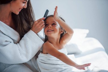 Tarak kullanarak saç stili yapmak. Genç anne kızıyla birlikte beyaz odada güzel bir gün geçiriyor..