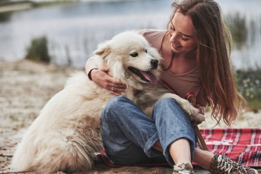 Arka plan bulanık. Güzel beyaz köpeğiyle sarışın kız kumsalda harika vakit geçiriyor..