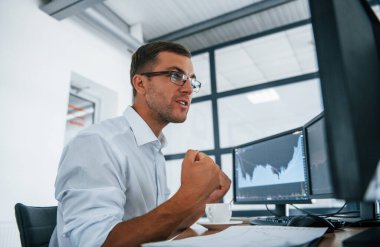 Heyecanlı ve mutlu. Zafer ve başarı kavramı. Resmi giyinmiş genç işadamı birden fazla ekranla ofiste..