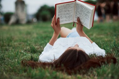 Kırmızı kapaklı kitap. Kız yeşil çimlere uzanıp dinleniyor..