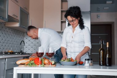 Her şey bitmek üzere. Beyaz gömlekli adam ve karısı mutfakta sebze kullanarak yemek hazırlıyorlar..