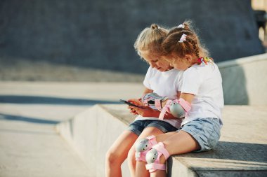 Akıllı telefon kullanıyor. Ekstrem sporlar için rampada. Dışarıda patenli iki küçük kız eğleniyor..