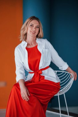 Kutlamaya dakikalar kaldı. Şenlikli kırmızı elbiseli güzel sarışın turuncu ve mavi renkli duvarları ve beyaz sandalyesi olan odada..