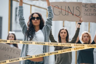 Kelime tabancalarının üzeri çizilmiş. Bir grup feminist kadın dışarıda hakları için protesto yapıyor..