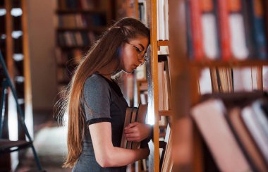 Raflara yaslanarak. Yorgun hissediyorum. Kütüphanede kitaplarla dolu bir kız öğrenci var. Eğitim kavramı.
