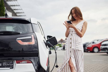 Beklerken akıllı telefon kullanmak. Elektrikli arabalardaki kadın gündüz şarj istasyonunda. Yepyeni bir araç..
