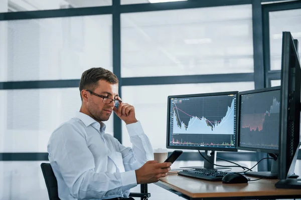 Akıllı telefon kullanıyor. Resmi giyinmiş genç iş adamı birden fazla ekranla ofiste. Değişim ve para kavramı.