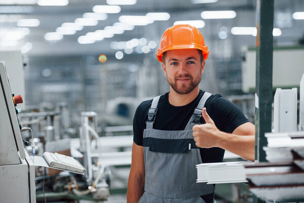 Shows thumb up. Well done. Everything is good. Industrial worker indoors in factory. Young technician with orange hard hat.