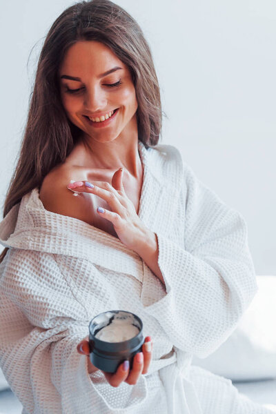 Bodycare by using cream. Portrait of beautiful brunette in white clothes that sits on the bed in room. Nice lighting.