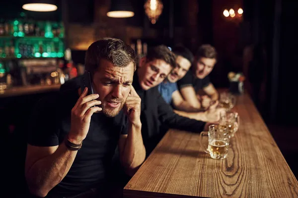 Adam telefonda konuşuyor. Bir barda futbol izleyen üç spor taraftarı. Ellerinde birayla.