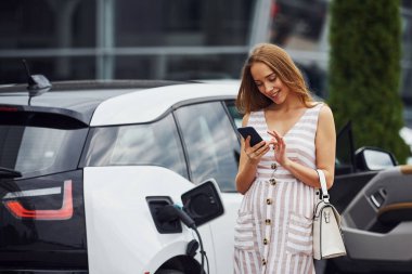 Şarj beklerken telefonu kullanmak. Gündüz vakti istasyonda bir kadın. Yepyeni bir araç..