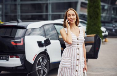 Şarj beklerken telefonu kullanmak. Gündüz vakti istasyonda bir kadın. Yepyeni bir araç..