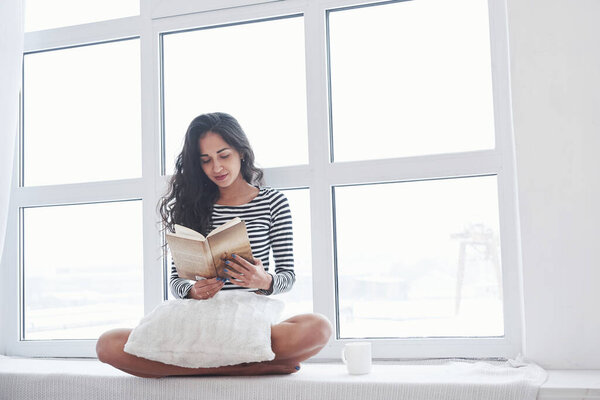 Girl reading book while sitting on the windowsill. Having free day.