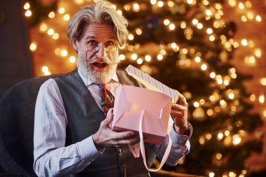 Gri saçlı ve sakallı son sınıf öğrencisinin portresi hediye kutusuyla süslenmiş Noel odasında..
