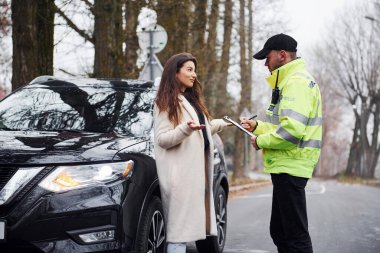Yeşil üniformalı erkek polis yolda arabanın kadın sahibiyle konuşuyor..