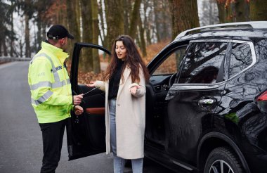 Yeşil üniformalı erkek polis yolda arabanın kadın sahibiyle konuşuyor..
