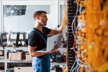 Üniformalı ve dizüstü bilgisayarlı genç adam sunucu odasında internet ekipmanı ve kablolarla çalışıyor..