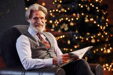 Gri saçlı ve sakallı son sınıf öğrencisinin portresi elinde kitapla süslenmiş Noel odasında..