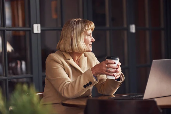 現代のラップトップと手にドリンクのカップとカフェ内に座っているシニア女性の肖像画 — ストック写真