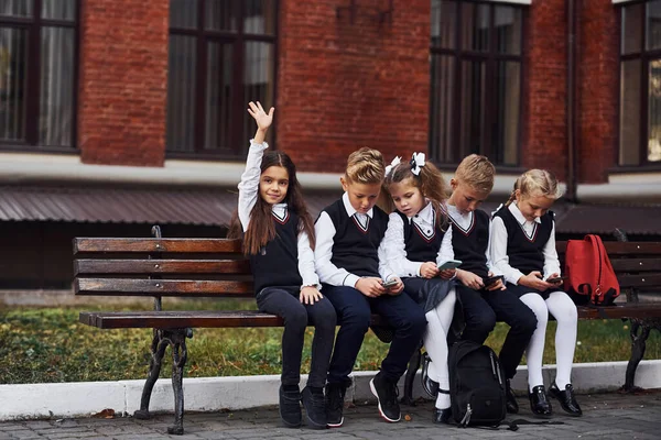 Grupp Barn Skoluniform Sitter Bänken Utomhus Tillsammans Nära Utbildningsbyggnad — Stockfoto