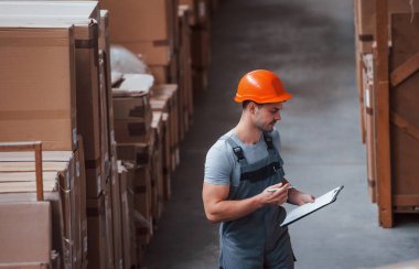 Üniformalı bir depo işçisi ve eldeki not defteri üretimi kontrol ediyor..