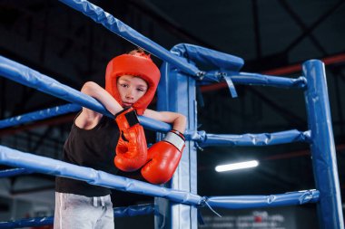 Boks ringinin düğümlerine dayanan koruyucu ekipmanlar içinde yorgun bir çocuk..