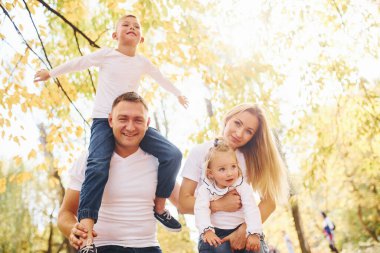 Anne ve baba çocukları omuzlarında ve ellerinde tutuyor. Neşeli genç aile birlikte bir sonbahar parkında yürüyüş yapıyor..
