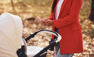 Elinde telefonla hem de. Kırmızı ceketli anne, sonbahar zamanı çocuğuyla parkta yürüyüşe çıkar..