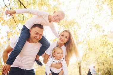 Anne ve baba çocukları omuzlarında ve ellerinde tutuyor. Neşeli genç aile birlikte bir sonbahar parkında yürüyüş yapıyor..
