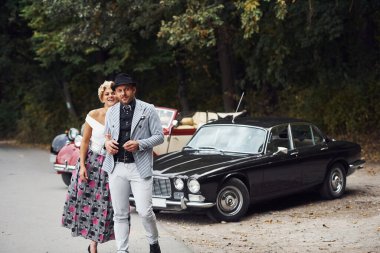 Eski moda güzel bir çift, arka planda orman olan eski model bir arabanın yanında giyiniyor..