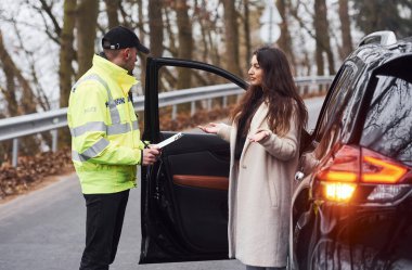 Yeşil üniformalı erkek polis yolda arabanın kadın sahibiyle konuşuyor..